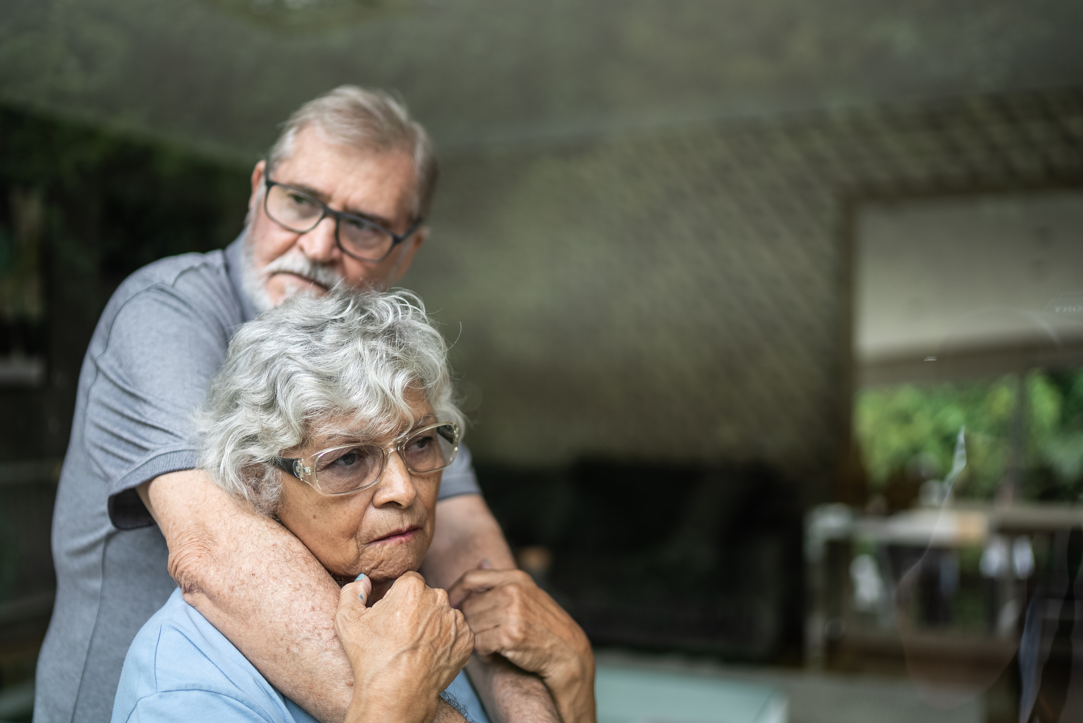 Two older adults appear worried, the man embraces the woman from behind.