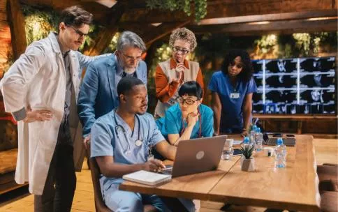 Medical professionals gathered around a table looking at a laptop screen, with medical scans displayed on a large monitor in the background.